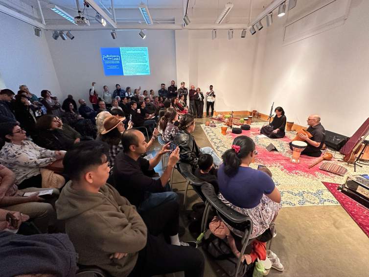 On the right side of the photo, Raheleh and Reza Filsoofi, two Iranian-American people, sit on a Persian carpet, surrounded by drums and other instruments.  On the left is a diverse audience watching the performance, some using their photos to record.