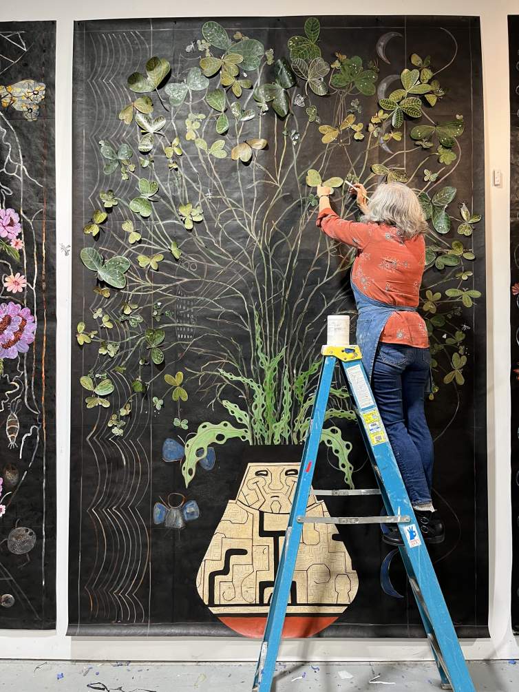 The artist stands on the third rung of a ladder and applies pale green leaves to a semi-glossy black sheet that stretches nearly ceiling to floor. Oval-shaped leaves splay like butterfly wings on the end of tall stems, which issue from a teardrop-shaped vase at the bottom center.
