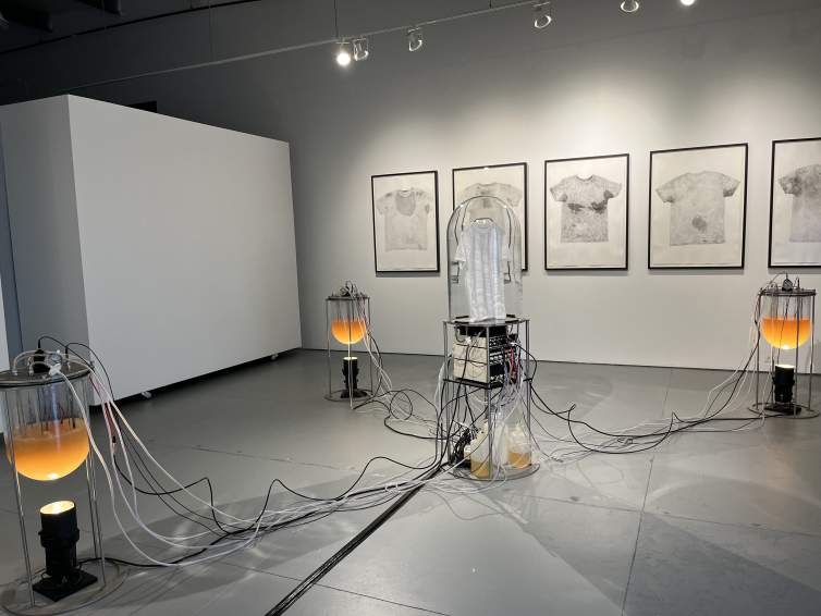 Installation view of three glass vats half-filled with yellow liquid and held in metal frames over spotlights like modern cauldrons. At each station, wires dip into the liquid and stretch to a central structure holding boxy machines that resemble stereo components. A white t-shirt is hung under a domed glass, and five framed shirts, each slightly stained with gray, hang across the wall opposite us.