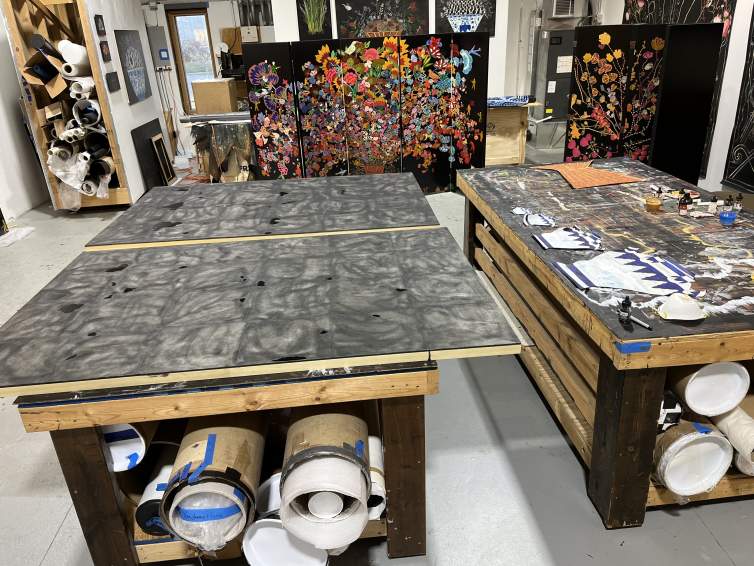 Two wood stretchers, covered with frosty-black material, overhang both ends of a work table in a studio. Inks and other materials sit on an adjacent table, and vividly colored floral compositions stand, nearly person-high, leaning against the far wall.