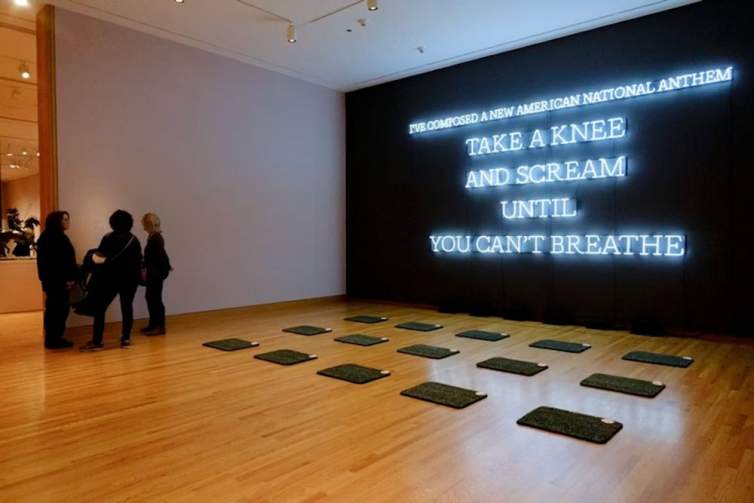 Neon American Anthem is installed in a gallery space, and is a black wall with white neon in block letters reading “I’ve composed a new american national anthem. Take a knee and scream until you can’t breathe”. In front of the neon are 15 rectangular kneeling pads.