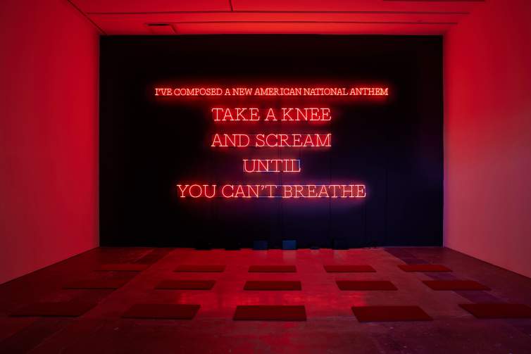 Neon American Anthem (red) is installed in a gallery space, and is a black wall with red neon in block letters reading “I’ve composed a new american national anthem. Take a knee and scream until you can’t breathe”. In front of the neon are 15 rectangular kneeling pads. The surrounding white walls are bathed in red light.