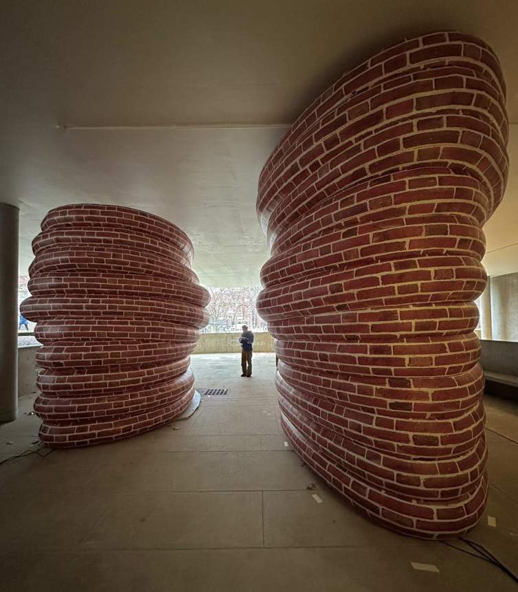 Jacqueline Kiyomi Gork stands at a distance between two huge oblong pillars make from coiled tubes painted with a brick pattern.