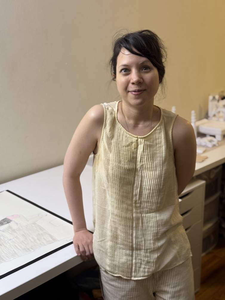 Emilie Louise Gossiaux leans against a drawing table in her home studio, smiling slightly at the camera. A petite woman of European and Indonesian descent, Gossiaux has dark brown hair pulled back in a messy bun, light tan skin, and wears a gauzy off-white tank top.