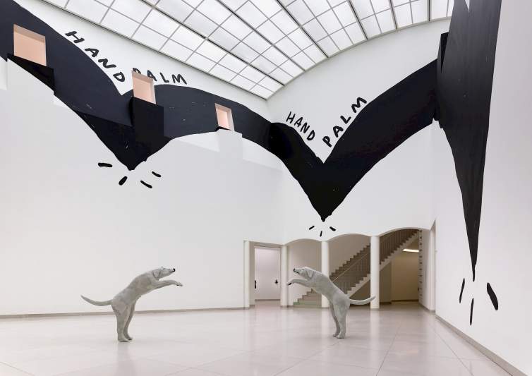 In a large gallery with white walls and floors, glass ceiling, and a black painted installation on the walls, two larger than life gray sculptures of an English Labrador Retriever standing upright on their hind legs. They are facing each other and have their front legs extended out in front of their bodies as though they are leaping into the air towards each other.