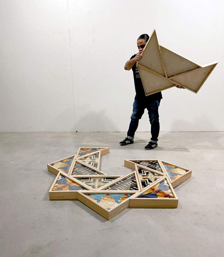 The artist Joe Harjo assembles pieces of a sunburst shaped artwork on the floor of a studio with white walls and concrete floors.