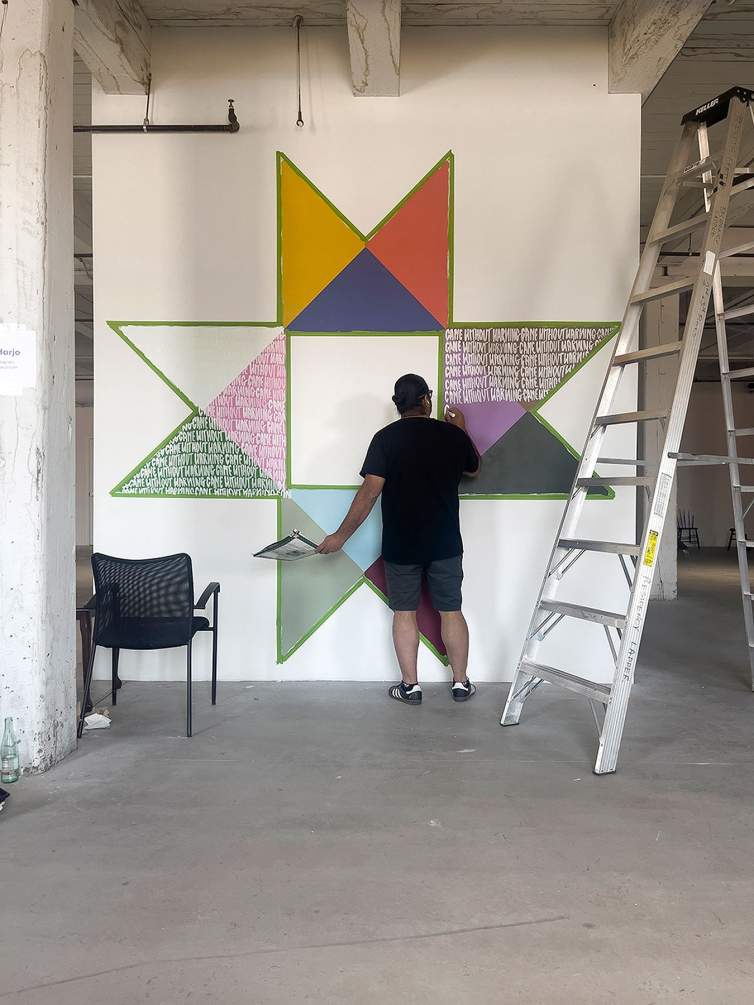In a studio with white walls and concrete floors, the artist Joe Harjo writes letters with white chalk on an artwork in progress,