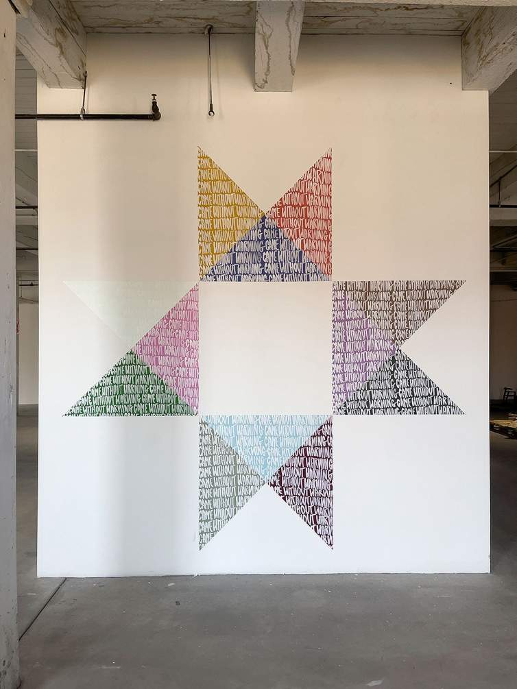 Came Without Warning is an artwork in a sunburst pattern painted on the wall. On the brightly colored triangles that make up the sunburst shape, the artist has written the words CAME WITHOUT WARNING in repeated lines.