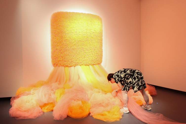 Ana Maria Hernando leans over a large sculpture, created from bundles of orange and pink tulle fabric, that flows down and out from a white wall in a gallery space with gray floors. Working with a bundle of fabric on the ground, she wears a patterned black and white dress over pink leggings and pink sneakers.