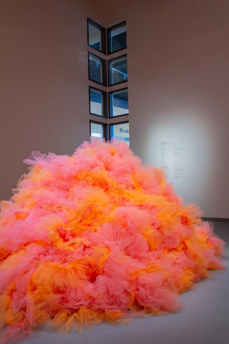 Making a Mountain is a large pile of bunched sections of orange and pink tulle fabric, installed in an art museum with gray floor, white walls, and tall windows visible in the corner.