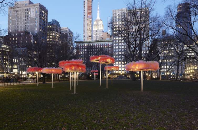 A twilight view of an urban park, with Ana María Hernando’s art installation “To Let the Sky Know.” We see unique, colorful sculptures resembling large mushrooms, against a backdrop of city buildings.