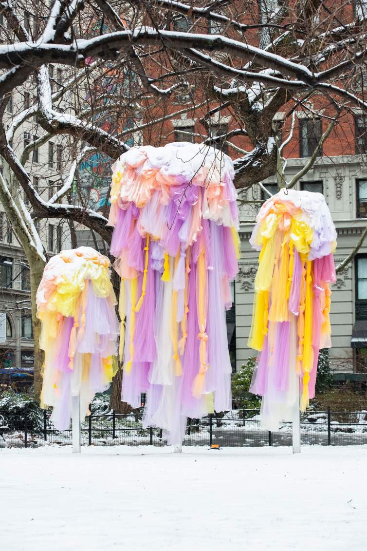 In an urban park with snow on the ground and buildings and trees in the background, Ana Maria Hernando’s sculptural installation, A Spring of Wild Kindnesses, features colorful tutu-like fabric art dusted with snow.