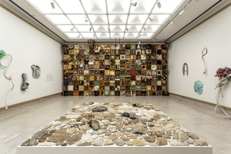 In a white walled gallery space are several installations. In the foreground is a shallow and wide square platform covered with  various flat stones in various textures and tones of taupe, grey and white. On the far back wall, a grid of 200 wood frames fill the wall top to bottom, each filled with a different object or assemblage.