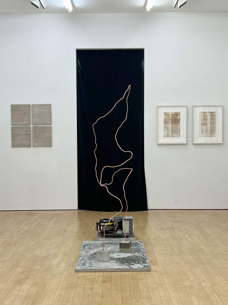 In an art gallery with wooden floors and white walls, a mechanical sculpture made of coiled copper refrigeration tubing sits on a concrete base in front of a dark door opening. Other abstract artworks hang on the walls.
