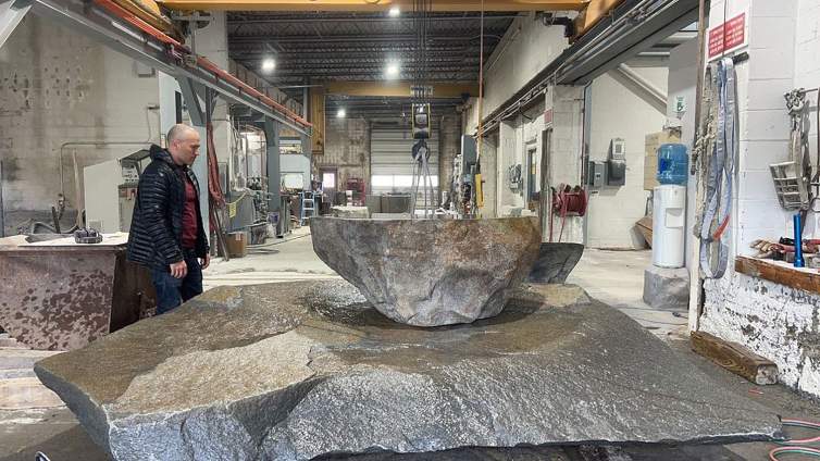The artist William Lamson, a Latino man with light skin tone and shaved black hair, stands in front of a large stone sculpture in progress in a large warehouse building.