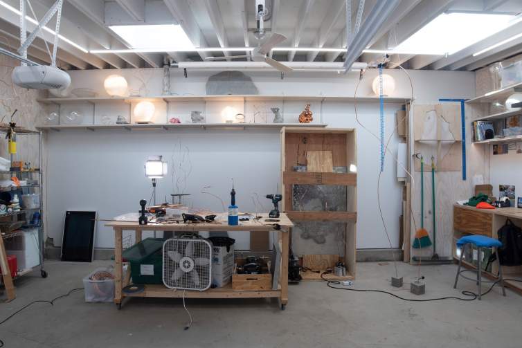 In an artist's studio with concrete floors, white walls and ceiling, we see a work table with multiple tools for woodworking and sculpture, along with lights and other household objects.