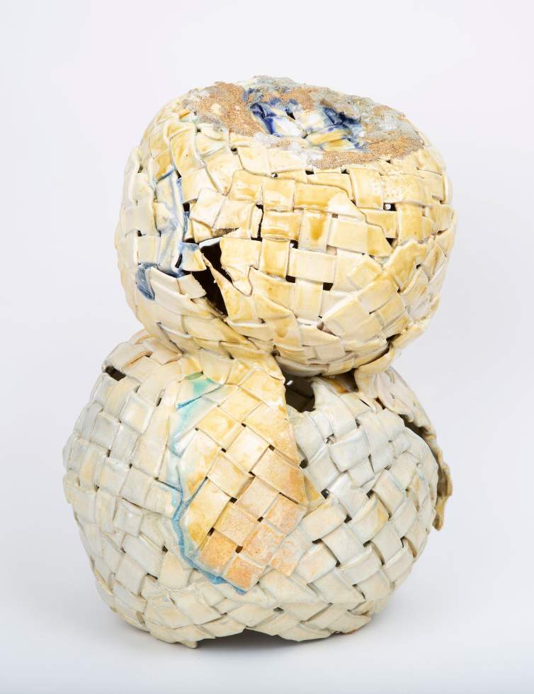 “Double Dutch” is a stoneware sculpture, with two stacked rounded forms structured of basket-like woven strips. The base form is slightly larger, and the top form leans forward. The glaze is off white and amber, with blue drips in places, and a crust of sand-like texture at the crown of the top form.