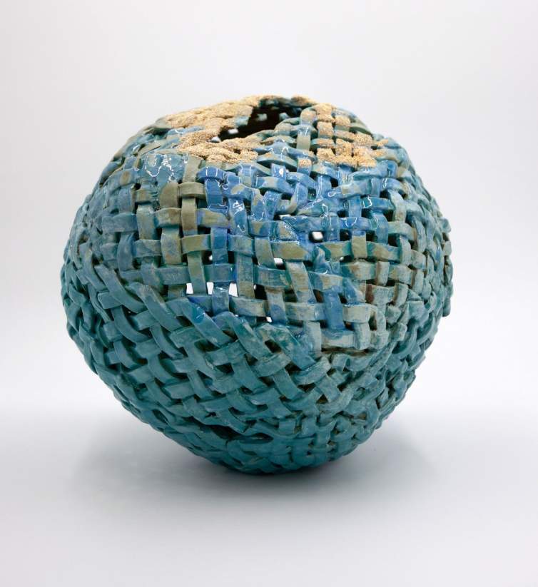 “Bubble” is a stoneware sculpture with a rounded shape, structured of basket-like woven strips. It is finished with a blue glaze with sand sprinkled over it.