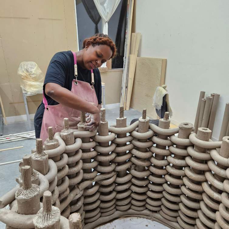 Anina Major, a Black woman with medium-dark skin tone and brown and black hair in braids, works with coils of clay to create a large sculptural woven form in a studio setting.