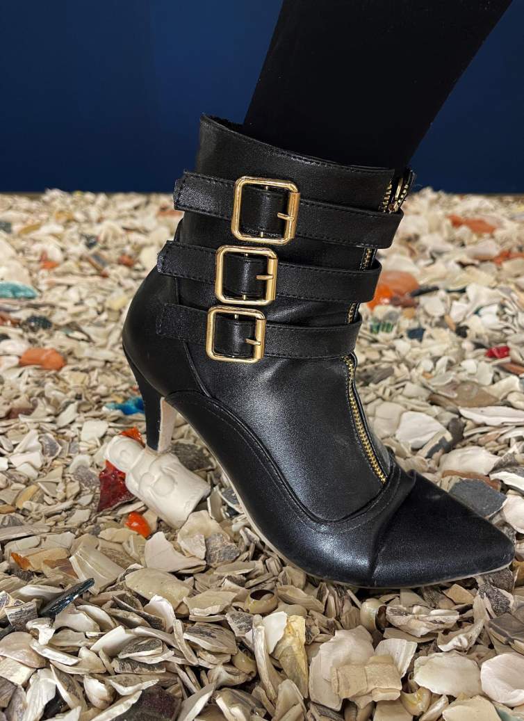 A black leather ankle boot with stiletto heel and three gold buckle straps stomps on a pile of ceramic shards and shells.