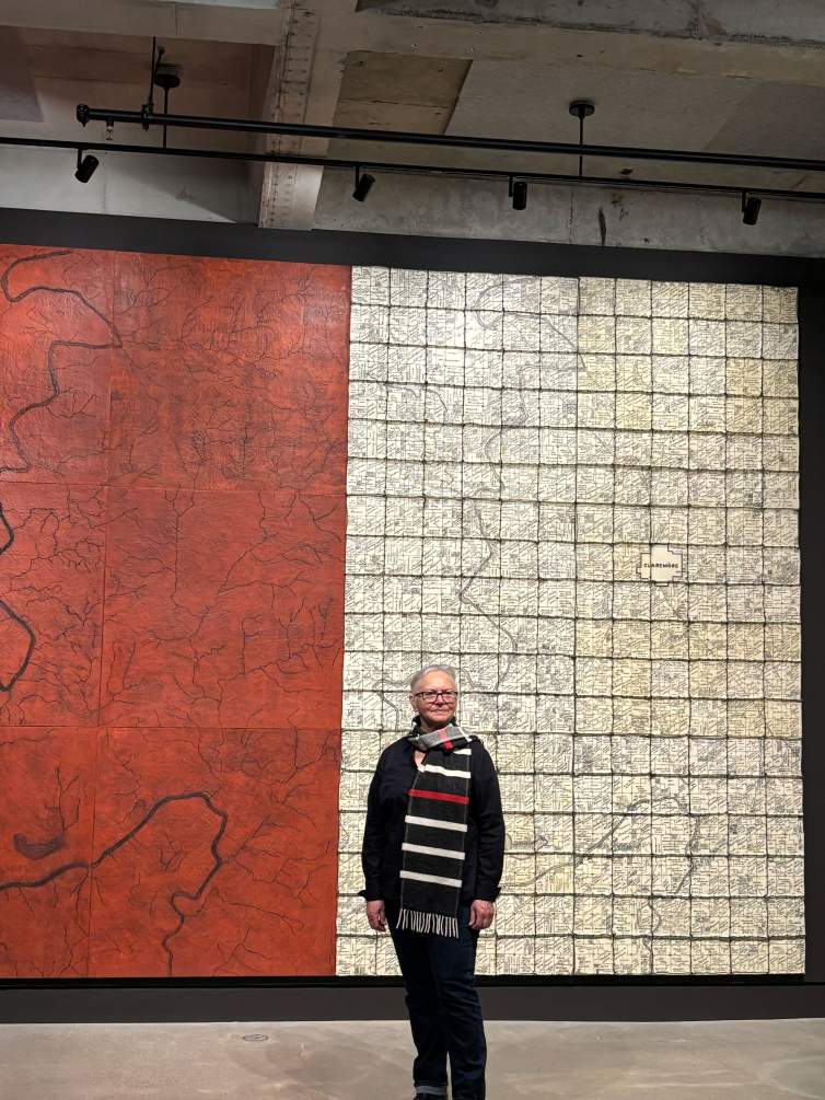 Brenda Mallory stands in front of a large abstract diptych. She is a citizen of Cherokee Nation, with long gray hair pulled back, light skin tone and wearing all black with a wide white and red striped black scarf.