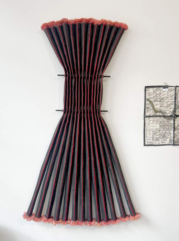 Thirteen equally long strips of black fire hose are bunched tightly together in the top half of the sculpture by a metal frame that hangs on a wall. Where they meet in the frame resembles pleating. Above and below the frame, the hose sections flare out slightly to make an hourglass shape. Each section is red down its front where the hose was flattened, and the frayed ends create red fuzz across the top and bottom edges.