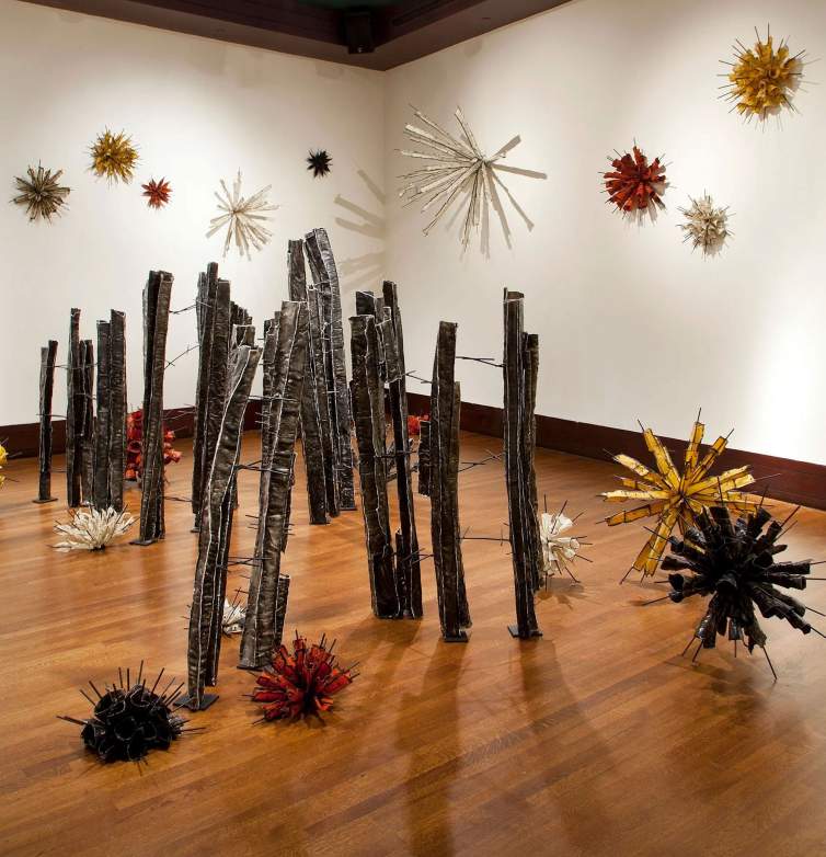 Installation view of a work made up of about 30 vertical, metal columns, some of which lean slightly and are connected with each other for barb-like rungs. Each column has a square or L-shaped cross-section. Eighteen sculptural asterisk shapes sit on the floor and hang on the wall like angular pompoms.