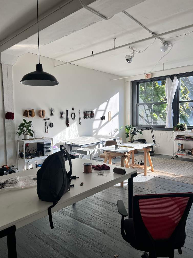 A backpack perches near the edge of a white work table in a white room lit only with sunlight pouring through windows on the wall to our right. More work surfaces are set up near the windows.