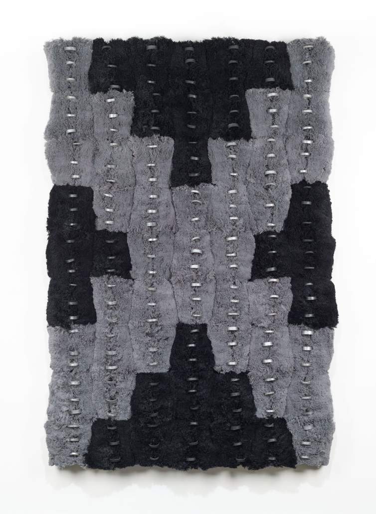 This vertically oriented panel is made up of strips of what first appears to be black textured fabric to all four sides of nickel-gray strips that together make a chunky X. Each strip is lined down its center with thin, horizontally oriented silver rings. A closer look reveals that the texture is created by the ends of countless cut threads.