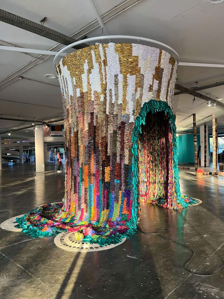 A columnar, hut-like form is made out of woven fabric that hangs from the edges of a disk at the ceiling of a deep, cavernous space. The textile is woven with gold, white, and silver around the top third or so, and it becomes more colorful as it moves down. The ends pool on the floor, and teal-blue fringe lines the arched opening and bottom edges of the textile.