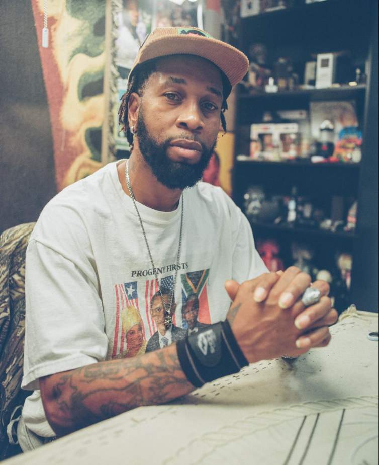 Demond Melancon is seated at a table in an interior space, facing the camera with hands clasped in front of him, resting on the table. He is a Black man with medium dark skin tone, light brown eyes, and a dark short beard and short dreadlocks. He wears a tan corduroy baseball hat, a silver chain with a medallion and a t-shirt with a faded graphic reading “Progeny Firsts” with photos of Sirleaf, Obama, and Mandela.