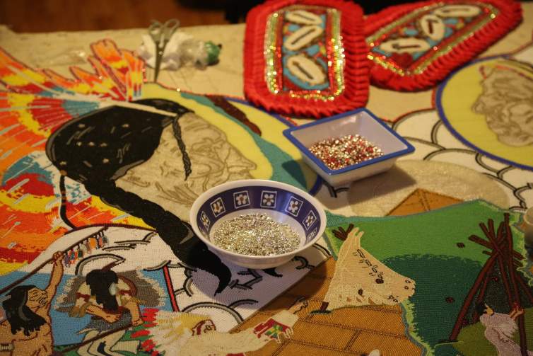 On a table is a narrative artwork created with small beads sewn onto canvas. Two small bowls of beads sit on the artwork, along with some tools and other beaded medallions. The scene in the sewn artwork depicts scenes interpreting stories about the Native American figures Red Cloud and Sitting Bull.