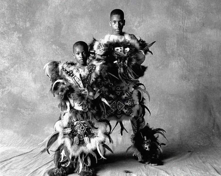 In a black-and-white portrait, two Black boys standing in front of a mottled fabric backdrop. Staring intently at the camera, they are dressed in Mardi Gras Indian costumes with abundant features and ornate beadwork.
