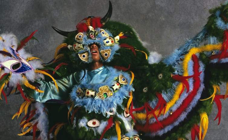 A photo of Demond Melancon performing in a brightly colored Mardi Gras Indian suit, adorned with feathers, boas, and beaded medallions, with horns on the headdress.