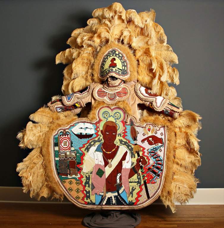 A photo of a human-sized Mardi Gras Indian costume adorned with golden yellow feathers and beaded medallions, including a large narrative beaded apron entitled “Bras-Coupé. The beaded artwork on the apron depicts an enslaved African prince who was known for leading rebellions against bondage. Bras-Coupé is pictured as a Black man holding a spear in one hand, and broken chains falling from the other amputated arm. Motifs flank the central figure including a throne, railroad tracks with flames, a sail ship, and a pointing hand holding a candle.