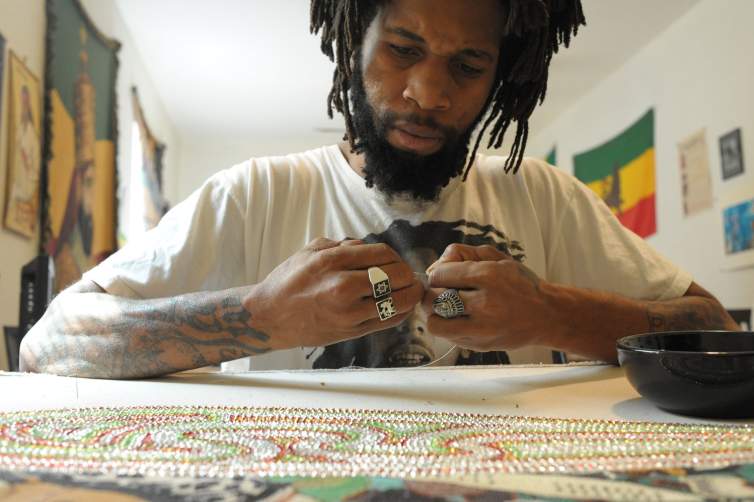 Demond Melancon, a Black man with medium-dark skin tone and black dreadlocks, is seated at a table working on a beaded sewing project.