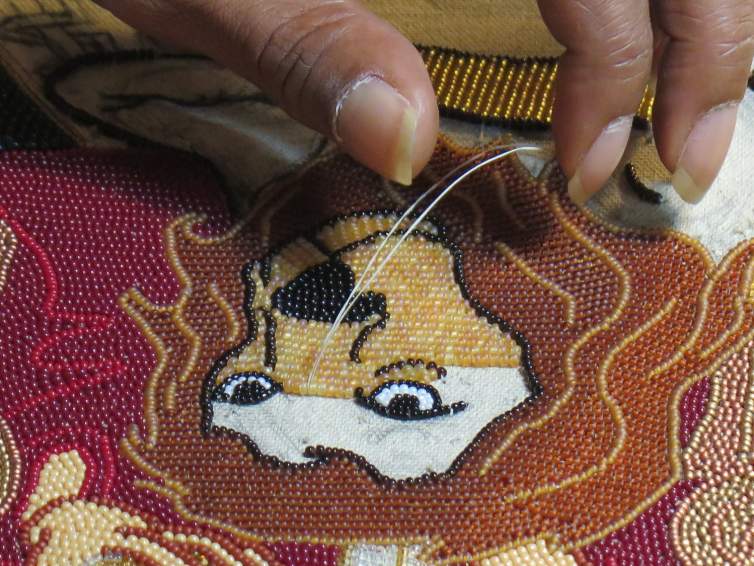 A close-up photo of a hand with medium-dark skin tone and long fingernails sewing tiny tiny colored beads to create a portrait of a lion.