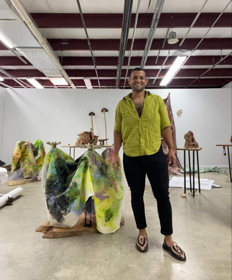 The artist Javier Orfon, a Puerto Rican person with medium skin tone, dark short hair, and face stubble, stands in a room with sculptural artworks behind him. He smiles at the camera, wearing a bright green shirt, left open at the top, black pants, and snakeskin loafers.
