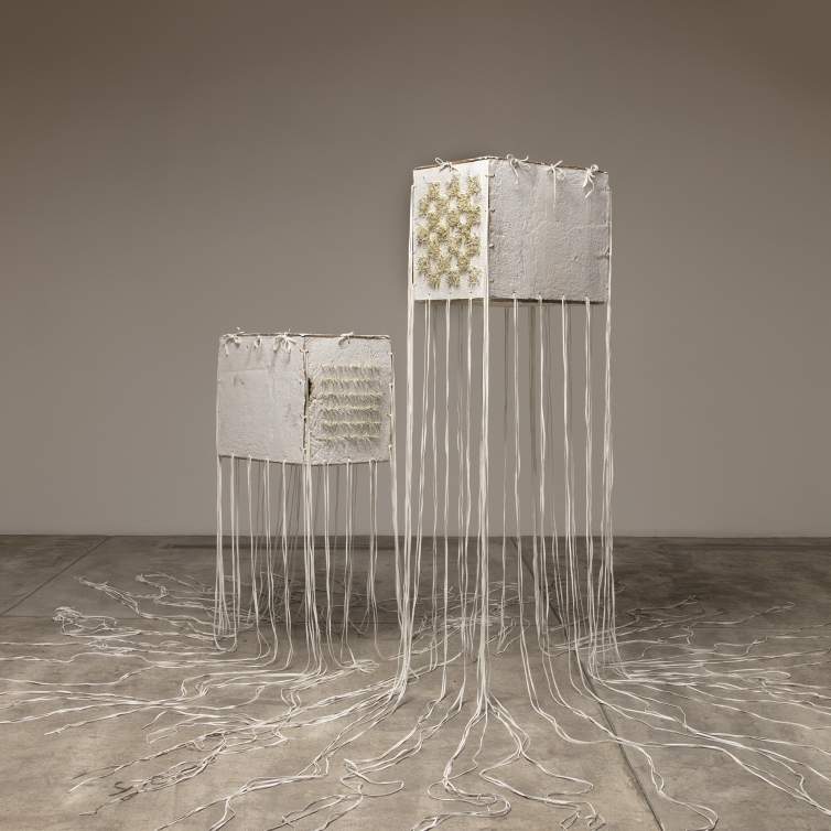 “Enduring” is a sculpture of 2 rectangular wide cubes made of textured off white handmade paper floating next to each other, bound at the edges with tied white deer hide laces. Long strings of lacing hang down from the edges and pool on the floor. The front cube is higher up in the air, and on one face has a checkerboard pattern of tan porcupine quills protruding an inch from the surface. The front face of the rear cube has 6 horizontal lines of quills protruding.
