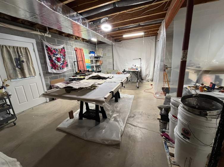 An artist’s studio in a basement with concrete floors, cinder-block walls, and natural wood ceiling beams. There is a central work table and a side area partitioned off with plastic sheeting.