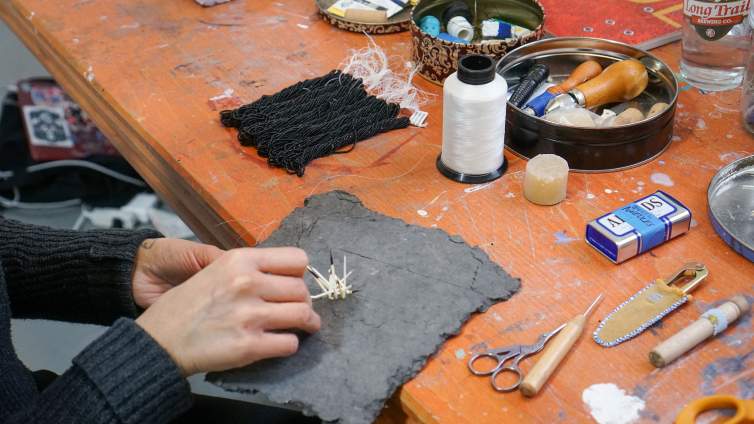 On an orange worktable, Mikayla Patton’s hands work with porcupine quills and dark handmade paper to create an element for a sculptural artwork. Various tools and materials are on the surface of the table.