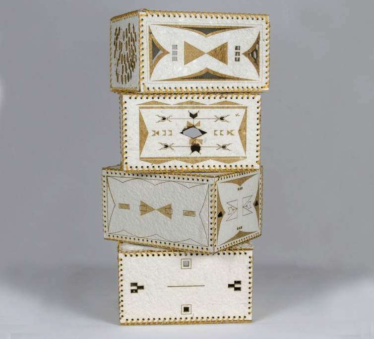 Anpa Kazanzan Win is a sculpture created with four stacked rectangular boxes, staggered at different angles. Each box is created with white handmade paper that is stitched together at the seams. The faces of the boxes are embellished with geometric patterns and abstract imagery.