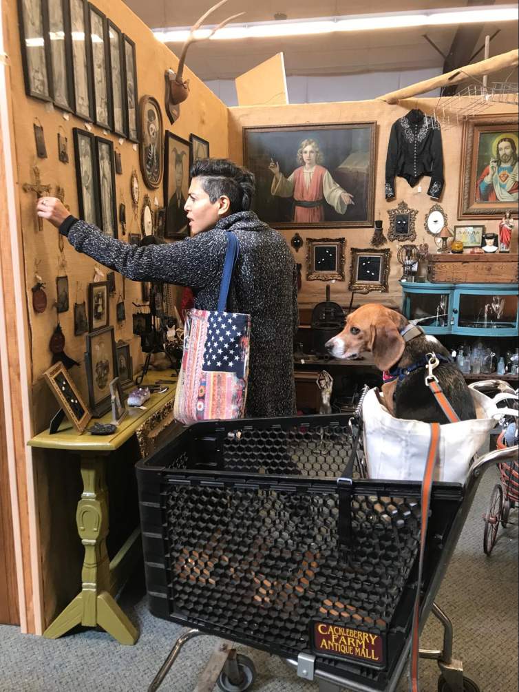 In a stall of an antiques mall, Sara reaches for a crucifix on a temporary wall densely hung with pictures and icons. Sara has light-toned skin and short black hair, and she wears a heathered gray sweater. A beagle sits in a bag in the basket of a shopping cart between us and the artist.