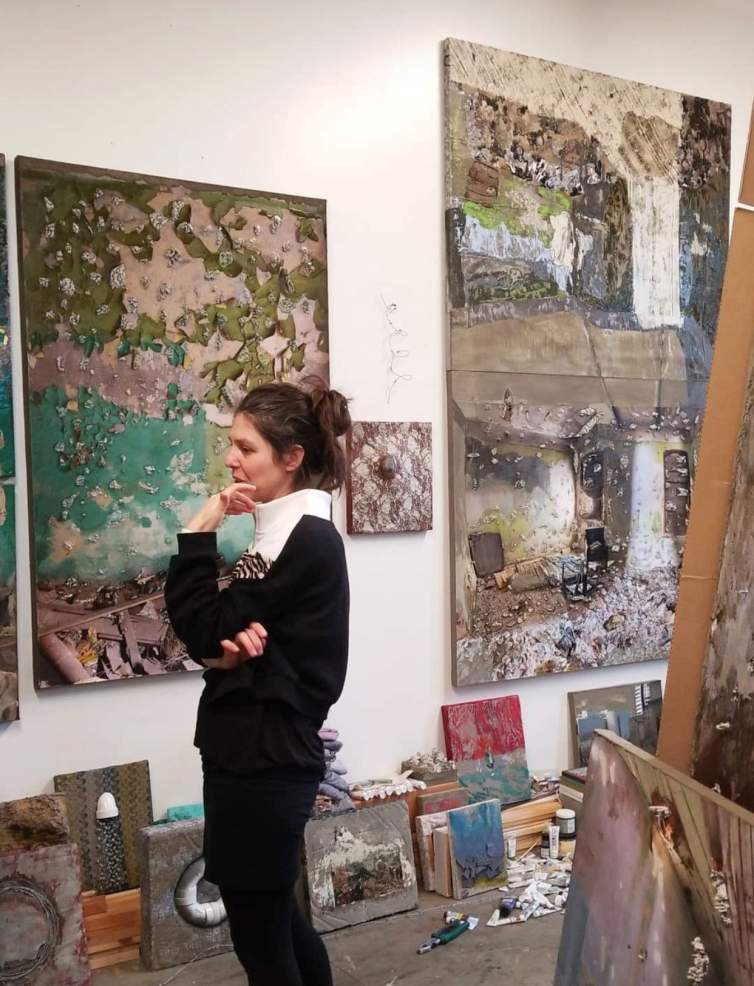 In an artist’s studio, a white woman with brown hair, a white turtleneck, and black sweater and pants touches her face with her hand while looking toward the left frame of the photo. On the wall are paintings of distressed interior spaces. Small paintings and art making materials surround the artist on the floor.