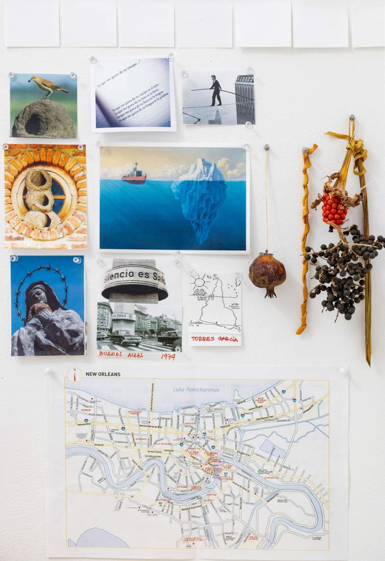 Various ephemera tacked to a studio wall above a table holding books, pens and pencils in cups. Papers on wall include blue color swatches, map of New Orleans, a blue abstract word form, and photograph of a tree.