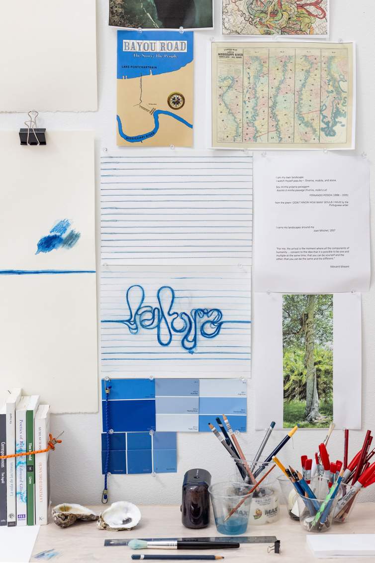 Various ephemera tacked to a studio wall above a table holding books, pens and pencils in cups. Papers on wall include blue color swatches, map of New Orleans, a blue abstract word form, and photograph of a tree.