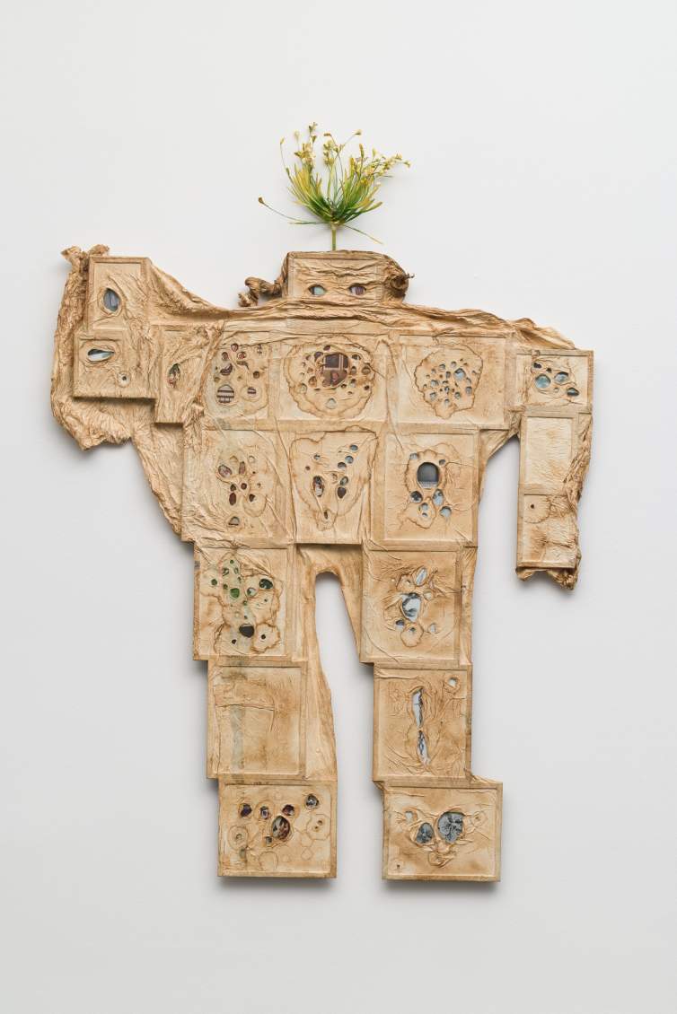 Picture frames are arranged flat and side by side to make a blocky human form and then overlaid with brown-stained, wrinkled paper. Glimpses of photos are visible through irregularly shaped holes piercing the paper, including a Nintendo Gameboy and a snapshot of a woman, though details on most are hard to make out.