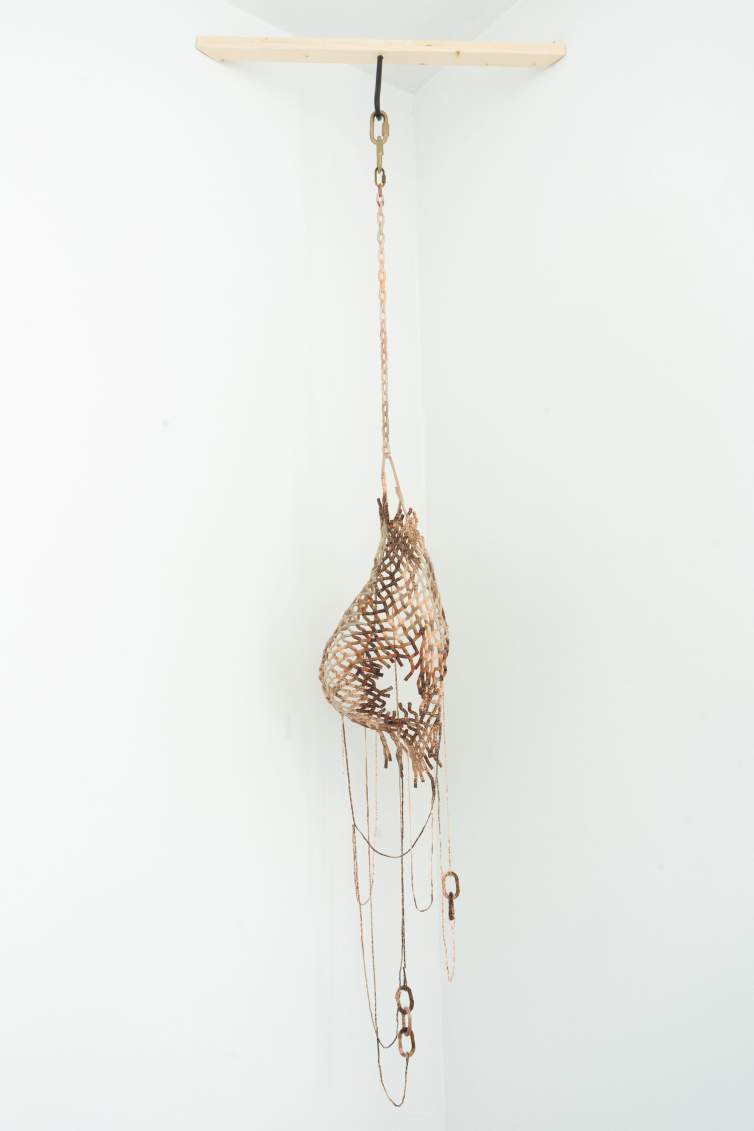 This sculptural piece hangs on a chain from a hook. A basket-like form curves in the shape of a breast. The strips are shades of brown, and they curl around a hole in the side facing us. Strands hang from the basket to loop and join with five chain links below.