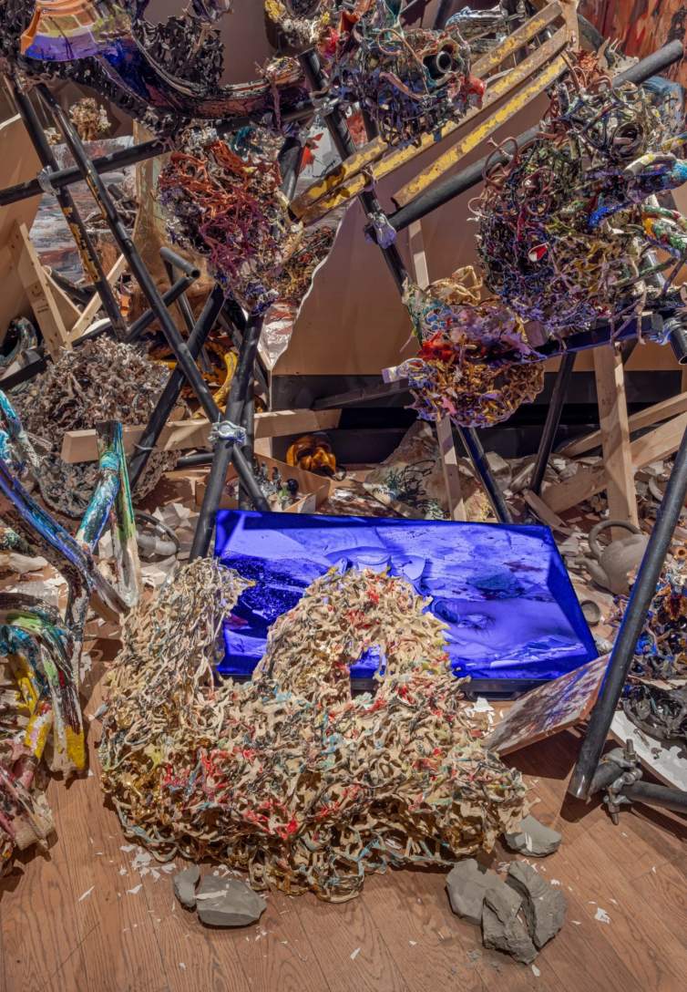 We look down onto a bright, indigo blue, roughly rectangular shape surrounded entirely by what appears to be confetti, shredded paper, and scraps. Some surfaces gleam, though, revealing that they are ceramic flecked with red, blue, green, and gold. That closer look also reveals a hand on part of the blue board.
