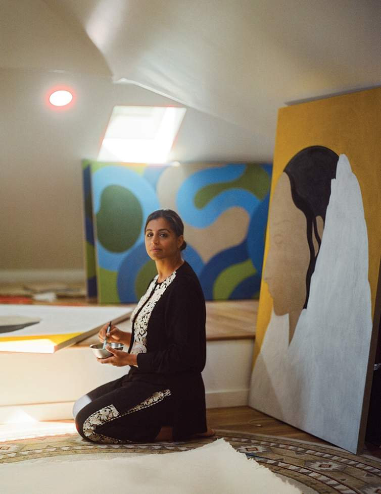 Rupy C. Tut kneels on the floor of a attic space with skylight and slanted ceilings , holding a bowl and paintbrush, surrounded by large paintings leaning on the wall and laying on the floor. She is wearing a matching black and floral set, and is an Indian-American woman with medium skin tone and long dark hair pulled into a bun.